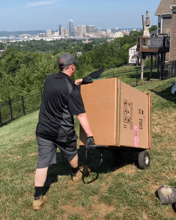 Logan Services HVAC technician bringing Trane furnace into customer's home with Cincinnati skyline in the background.